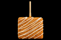 Square covered crispy treat with white drizzle on a stick against a black background.