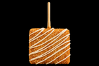 Square covered crispy treat with white drizzle on a stick against a black background.