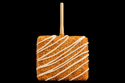 Square covered crispy treat with white drizzle on a stick against a black background.